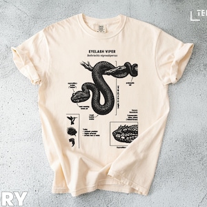 Może przedstawiać: Kremowa koszulka z czarną ilustracją grzechotnika rzęskowego, zawierającą szczegóły anatomiczne i napis "EYELASH VIPER". Koszulka ma okrągły dekolt i krótkie rękawy.