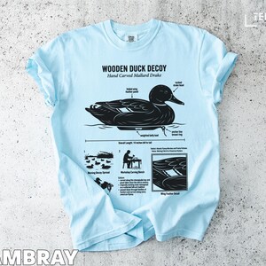 Light blue t-shirt with black graphic of a wooden mallard drake decoy and informational text.