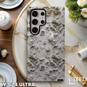 Puede incluir: Una funda de tel&eacute;fono blanca con un dise&ntilde;o en relieve 3D de flores y un fantasma. La funda est&aacute; dise&ntilde;ada para el Galaxy S24 Ultra.