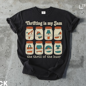 May include: Black t-shirt with the text "Thrifting is my Jam" and illustrations of jars containing vintage items. The phrase "the thrill of the hunt" is printed below the jars. The shirt is made of a soft material.