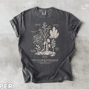 May include: Dark grey t-shirt featuring a botanical illustration with the text "THE SHADOW HERBARIUM". The design includes detailed drawings of plants and their parts, with labels. The shirt has a vintage, worn look.