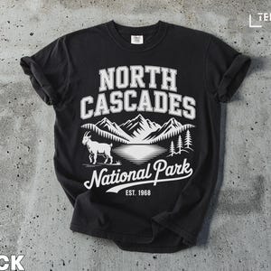 Pode incluir: Camiseta preta com um gráfico branco com as palavras "North Cascades National Park" e uma cena de montanha com uma cabra. O texto "EST. 1968" também está incluído no design.