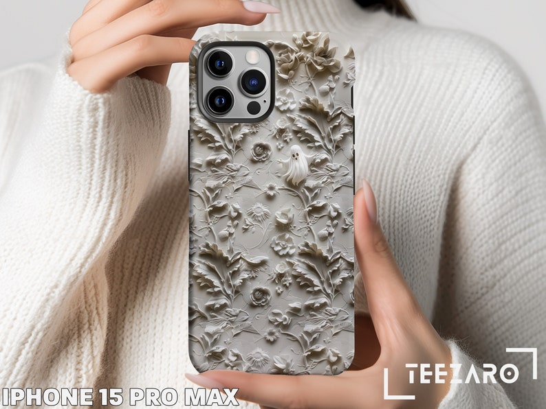 A person holds an iPhone 15 Pro Max with a case featuring an intricate monochromatic floral design with a subtle ghost hidden among the leaves. The delicate details contrast against the person's cozy white knit sweater, highlighting.