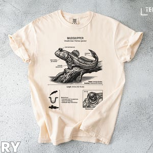 May include: Ivory-colored t-shirt featuring a detailed illustration of a mudskipper fish, with scientific annotations and diagrams. The graphic includes the text "MUDSKIPPER" and measurements of 4-6 inches (10-15 cm) in length. The shirt has a classic crew neck.