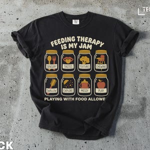 Puede incluir: Camiseta negra con el texto "FEEDING THERAPY IS MY JAM" e ilustraciones de tarros que contienen elementos relacionados con la comida. La camiseta también dice "PLAYING WITH FOOD ALLOWE".