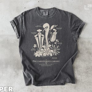 Puede incluir: Camiseta gris oscuro con una ilustración botánica de plantas carnívoras y etiquetas detalladas. El texto "THE CARNIVOROUS CABINET" está impreso debajo. La camiseta tiene mangas enrolladas y cuello redondo.