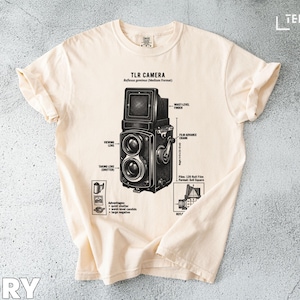 May include: Ivory-colored t-shirt featuring a detailed diagram of a TLR camera. The graphic includes labels for various camera parts, such as the viewing lens and film advance crank. The shirt's design is a vintage-inspired illustration, perfect for photography enthusiasts.
