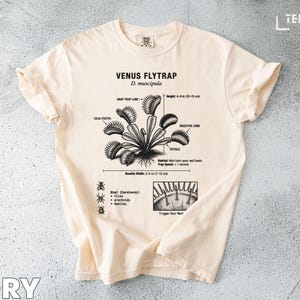 May include: Ivory-colored t-shirt featuring a detailed black and white illustration of a Venus Flytrap, with labeled parts and measurements in inches. The shirt includes the text "VENUS FLYTRAP D. muscipula".