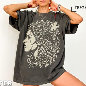 Może przedstawiać: Ciemnoszara koszulka z profilem kobiety i wyjącym wilkiem. Włosy kobiety i wilk są splecione z liściastym wzorem. Słowo "TEEZARO" jest wydrukowane w prawym górnym rogu.