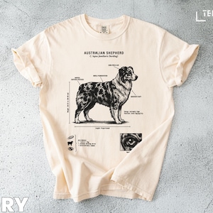 May include: Ivory-colored t-shirt featuring a detailed illustration of an Australian Shepherd dog with anatomical labels. The graphic includes the dog's measurements and a close-up of its eye. The text "Australian Shepherd" is printed at the top.
