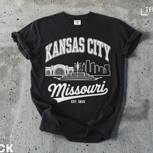 Può includere: T-shirt nero con un design grafico bianco con "Kansas City" in un arco, uno skyline e "Missouri" in corsivo. Il testo "EST. 1853" è in basso. La maglietta è su una superficie di cemento grigio.
