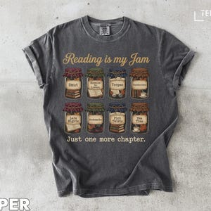 May include: Dark gray t-shirt with the text "Reading is my Jam" and illustrations of jars labeled with book-related terms like "Smut" and "Tropes." The phrase "Just one more chapter" is below the jars.
