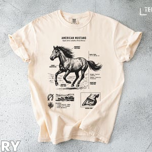 Könnte beinhalten: Elfenbeinfarbenes T-Shirt mit einer detaillierten Illustration eines American Mustang-Pferdes. Die Grafik enthält anatomische Details und den Text "AMERICAN MUSTANG". Das Shirt hat kurze Ärmel und einen Rundhalsausschnitt.