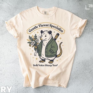 Puede incluir: Camiseta de color marfil con un dibujo de un opossum con una chaqueta verde y flores. El texto dice "Gentle Threat Specialist" encima del opossum y "Soft Voice Sharp Teeth" debajo. La camiseta tiene las mangas dobladas.