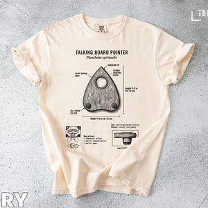 May include: Ivory t-shirt with a planchette pointer graphic, labelled with dimensions and text: "Talking Board Pointer" and "Planchette spiritualis". The planchette is 10-13 cm wide. The design includes a diagram of the planchette's parts.