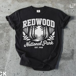 Pode incluir: Camiseta preta com um design gráfico branco do Parque Nacional Redwood. O design inclui o texto "REDWOOD" acima de uma cena florestal e "National Park EST. 1968" abaixo. A camiseta é feita de material macio.