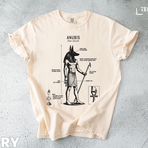 Puede incluir: Camiseta color marfil con una ilustración detallada de Anubis, el dios con cabeza de chacal del antiguo Egipto. El gráfico incluye detalles anatómicos y etiquetas de texto. La camiseta tiene mangas enrolladas y cuello redondo. Medidas en cm.