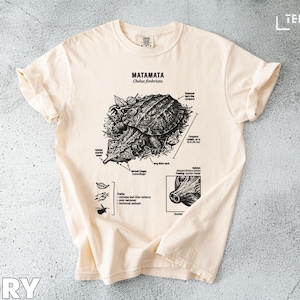 Puede incluir: Camiseta color marfil con una ilustración en blanco y negro de una tortuga Mata Mata y anotaciones científicas. Incluye el texto "MATAMATA Chelus fimbriata".