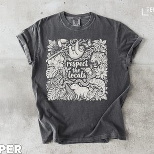 May include: A charcoal gray t-shirt featuring a white graphic print of various rainforest animals including a sloth, toucan, jaguar, and capybara, surrounded by tropical foliage. The text "respect the locals" is centered in the design.