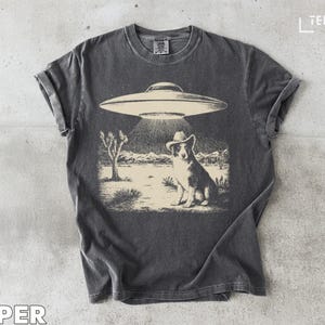 Puede incluir: Camiseta gris oscuro con un diseño gráfico de un perro con sombrero de vaquero iluminado por un OVNI, sobre un paisaje desértico. La palabra "PEPPER" está impresa en la parte inferior.