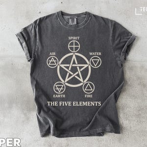 Peut inclure: T-shirt gris foncé avec un motif crème représentant un pentagramme et des symboles des cinq éléments : Esprit, Air, Eau, Terre et Feu. Le texte "THE FIVE ELEMENTS" est en dessous. Les manches sont retroussées.