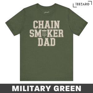 May include: A military green t-shirt with the text "Chain Smoker Dad" in a white vintage font. A disc golf disc is featured in the text.