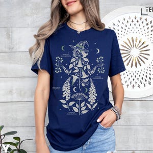 Puede incluir: Camiseta azul marino con una ilustraci&oacute;n bot&aacute;nica de una bruja, con detalles florales y lunares. El dise&ntilde;o incluye etiquetas de texto y est&aacute; sobre un fondo claro de vetas de madera. La camiseta tiene mangas cortas y cuello redondo.