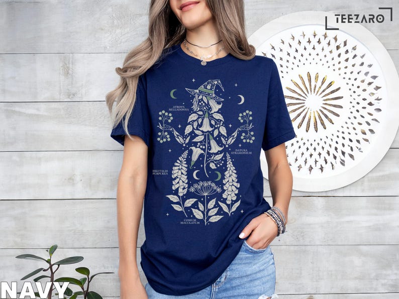 Puede incluir: Camiseta azul marino con una ilustraci&oacute;n bot&aacute;nica de una bruja, con detalles florales y lunares. El dise&ntilde;o incluye etiquetas de texto y est&aacute; sobre un fondo claro de vetas de madera. La camiseta tiene mangas cortas y cuello redondo.