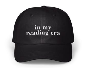 May include: Black baseball cap with the phrase "in my reading era" printed in white. The cap has a curved brim and a structured fit. This hat is a stylish accessory for book lovers.