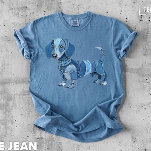 Puede incluir: Camiseta azul claro con un diseño de perro salchicha de retazos de mezclilla. El perro tiene orejas azules, un collar de mezclilla y un cuerpo de mezclilla. El texto "BLUE JEAN" está en la parte inferior de la camiseta.