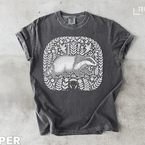 Può includere: T-shirt grigio scuro con un'illustrazione bianca di un tasso circondato da elementi floreali. Il tasso è sdraiato e il disegno è incorniciato da una ghirlanda di foglie e fiori. La parola "PEPPER" è in basso.