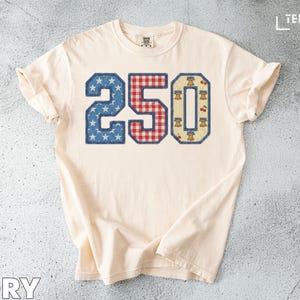 May include: Ivory-coloured t-shirt featuring the number 250 in a patriotic design. The number 2 is blue with white stars, the 5 is red and white checkered, and the 0 is beige with cherries and bells.