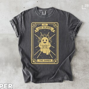 May include: Dark gray t-shirt featuring a gold-colored tarot card design. The card depicts a ghost holding a game controller, with the text "LAG SLAYER" and "THE GAMER". The shirt has a vintage, worn look.