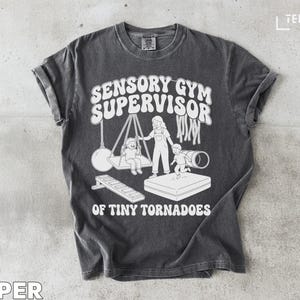 Può includere: T-shirt grigio scuro con la scritta bianca "SENSORY GYM SUPERVISOR OF TINY TORNADOES". La grafica raffigura una persona, bambini e attrezzature da palestra. La maglietta è realizzata in un materiale morbido e confortevole.