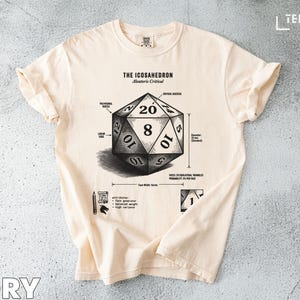 May include: Ivory-colored t-shirt featuring a detailed illustration of an icosahedron, a 20-sided die, with labels and the text "The Icosahedron Aleatoris Critical." The design includes numbers and diagrams, perfect for tabletop gaming enthusiasts.