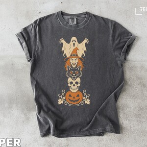 May include: A charcoal grey short-sleeve t-shirt featuring a vertical graphic stack of Halloween icons including a ghost, a witch, a black cat, a skull, and a jack-o'-lantern. The text TEEZARO is visible in the top right corner and PEPPER at the bottom.