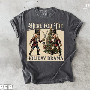 Puede incluir: Camiseta gris oscuro con un diseño gráfico vintage. Presenta dos soldados cascanueces con espadas, un árbol de Navidad y el texto "HERE FOR THE HOLIDAY DRAMA".