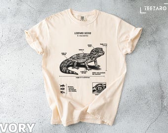 Może przedstawiać: Kremowa koszulka z czarno-białą ilustracją gekona lamparciego, z oznaczeniami anatomicznymi i napisem "LEOPARD GECKO". Długość gekona wynosi 20-28 cm. Koszulka zawiera również zbliżenie oka gekona.