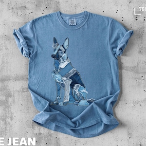 Puede incluir: Camiseta azul claro con un diseño de perro sentado hecho con parches de mezclilla. Las mangas están enrolladas. Las palabras "BLUE JEAN" están impresas en la parte inferior. La marca "TEEZARO" está impresa en la esquina superior derecha.