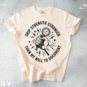 May include: Ivory-colored t-shirt with a black graphic. The graphic features a hand gripping a dynamometer, surrounded by text that reads "Grip Strength Stronger Than My Will To Document". The shirt has rolled sleeves.