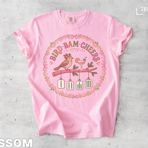 Pink t-shirt with "Bird Bam Cheers" text, a bird on a branch, and cocktails.