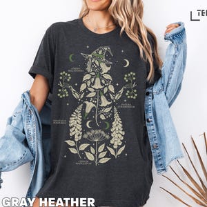 Puede incluir: Camiseta gris oscuro jaspeado con un dise&ntilde;o de bruja bot&aacute;nica. El dise&ntilde;o incluye una silueta de bruja con sombrero, lunas crecientes e ilustraciones de plantas con nombres cient&iacute;ficos. El texto "DARK GRAY HEATHER" est&aacute; en la parte inferior.