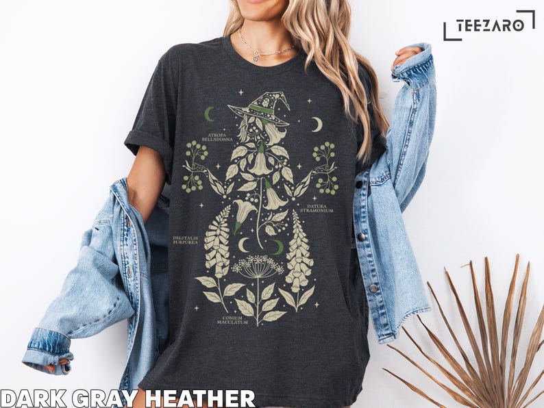 Puede incluir: Camiseta gris oscuro jaspeado con un dise&ntilde;o de bruja bot&aacute;nica. El dise&ntilde;o incluye una silueta de bruja con sombrero, lunas crecientes e ilustraciones de plantas con nombres cient&iacute;ficos. El texto "DARK GRAY HEATHER" est&aacute; en la parte inferior.