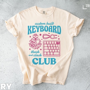 May include: Ivory t-shirt with a teal and pink graphic. The design includes the words "custom built KEYBOARD CLUB", keyboard component illustrations, and the text "thock and clack". The shirt is made of a soft material. The t-shirt is a neutral colour.
