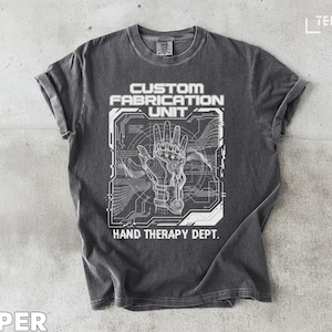 Peut inclure: T-shirt gris foncé avec un motif blanc d'une main robotique et le texte "CUSTOM FABRICATION UNIT" et "HAND THERAPY DEPT." Le design comprend des éléments de circuits imprimés. Le t-shirt est à col rond.