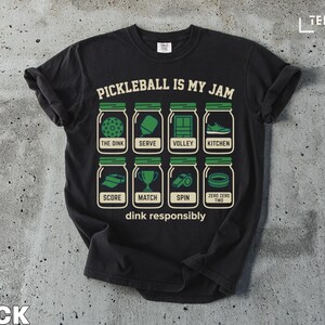 May include: Black t-shirt featuring the phrase "PICKLEBALL IS MY JAM" and images of pickleball equipment in jars. The shirt also has the text "dink responsibly".