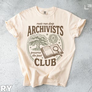 Puede incluir: Camiseta de color marfil con un diseño gráfico para el Club de Archivistas. El diseño incluye un árbol, un libro abierto con una lupa y las palabras "roots run deep" y "preserve the past".