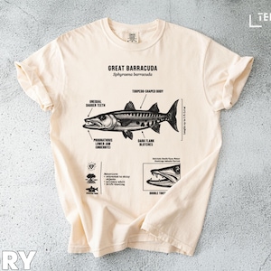 Puede incluir: Camiseta de color marfil con una ilustración detallada de una Gran Barracuda, que incluye detalles anatómicos y el texto "Great Barracuda". La camiseta tiene las mangas enrolladas y está colocada sobre una superficie texturizada.