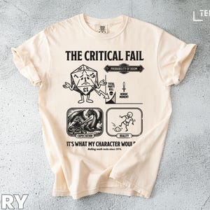 May include: Ivory-colored t-shirt with a black graphic design. The design includes the text "THE CRITICAL FAIL" and illustrations of a die, a dragon, and a stick figure. The text "IT'S WHAT MY CHARACTER WOUI" is also present.