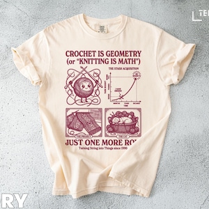 May include: Ivory-colored t-shirt with a maroon graphic. The graphic includes the text "CROCHET IS GEOMETRY (or "KNITTING IS MATH")", a yarn ball character, knitting needles, and a graph. The shirt also has the text "JUST ONE MORE RO" and "Turning String into Things since 1990".